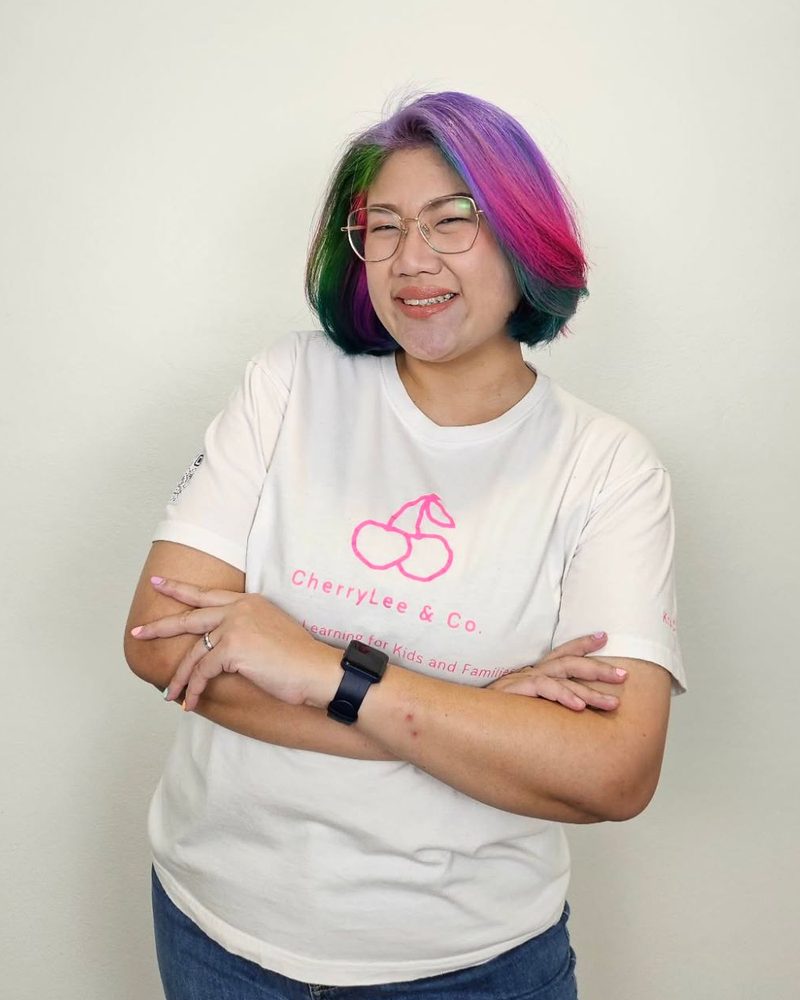 ครูเชอร์รี่ ผมสีรุ้ง สวมแว่น ยิ้มกอดอกในเสื้อยืดสีขาวลายโลโก้ CherryLee & Co.